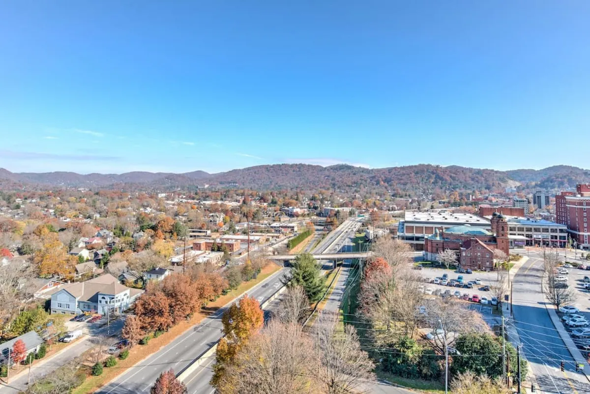 A sweeping elevated view over Asheville, featuring rolling hills, the town spread below, a meandering highway, and an abundance of colorful autumn foliage. The landscape evokes a sense of exploration and invites guests to discover both the urban and natural aspects of the area.