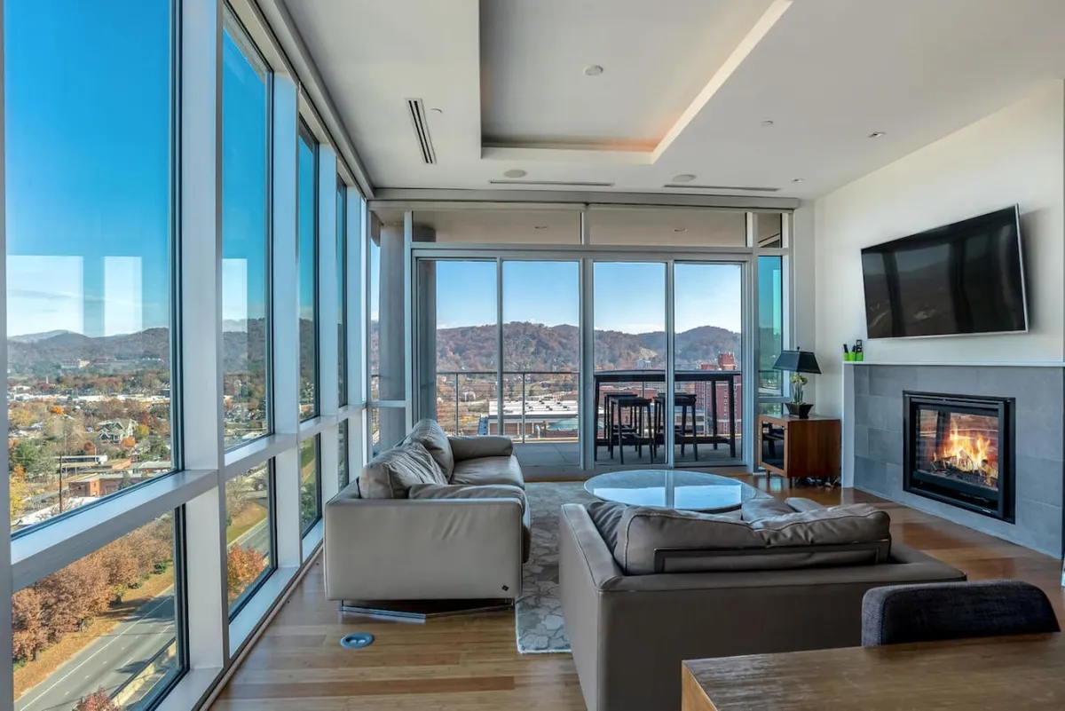 The image showcases an inviting, contemporary living area with floor-to-ceiling windows framing sweeping views of the Blue Ridge Mountains. A modern gas fireplace adds warmth and ambiance, while plush seating creates a perfect spot to unwind. In the background, a dining table promises memorable meals shared with family or friends, all set against the stunning mountain backdrop.