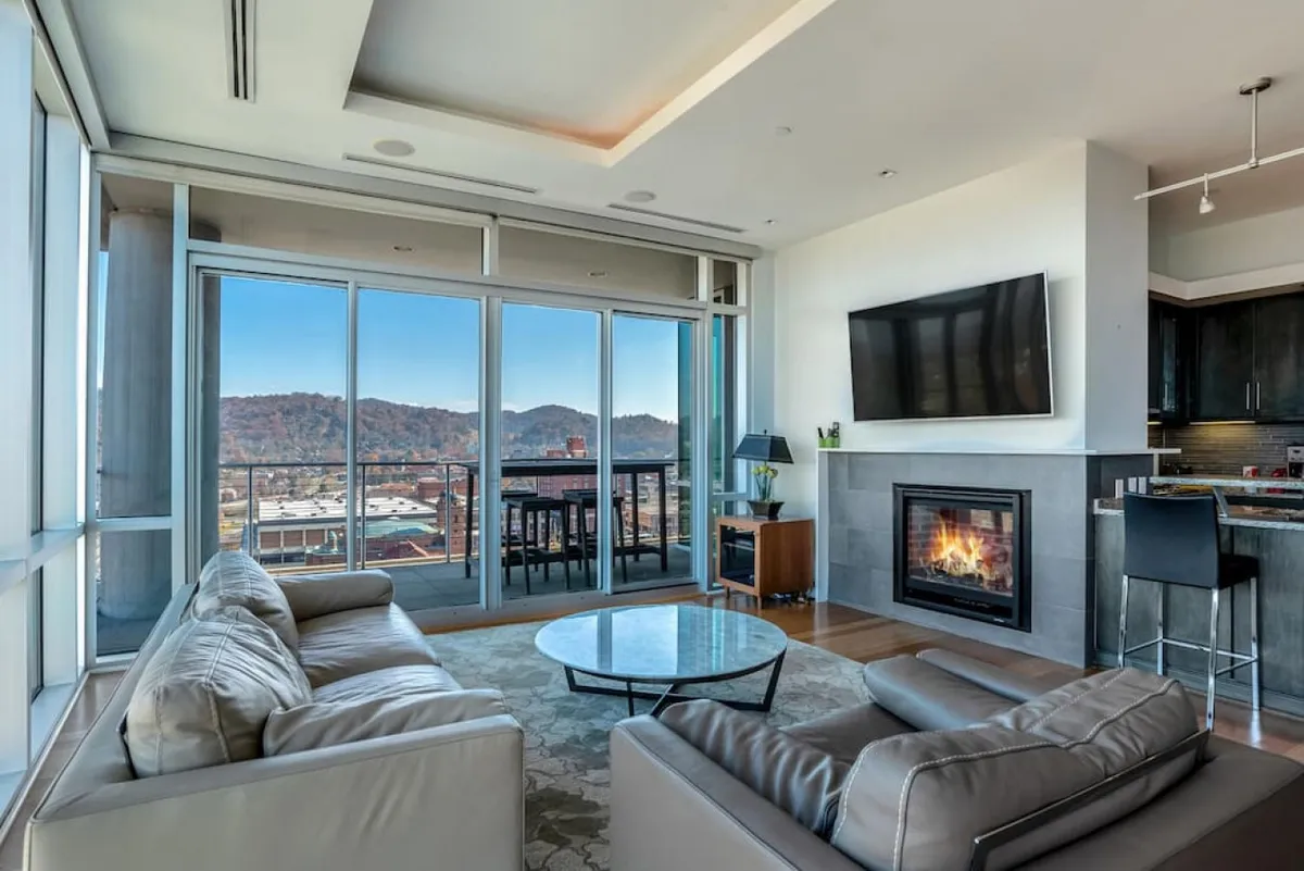 The image showcases an inviting living room bathed in natural light from expansive floor-to-ceiling windows that frame panoramic views of the Blue Ridge Mountains. Plush seating is arranged around a sleek, contemporary gas fireplace, with a coffee table and TV enhancing the space’s comfort. The hardwood floors and subtle modern decor add refinement, while the sunrise glow outside hints at the serenity and beauty beyond.