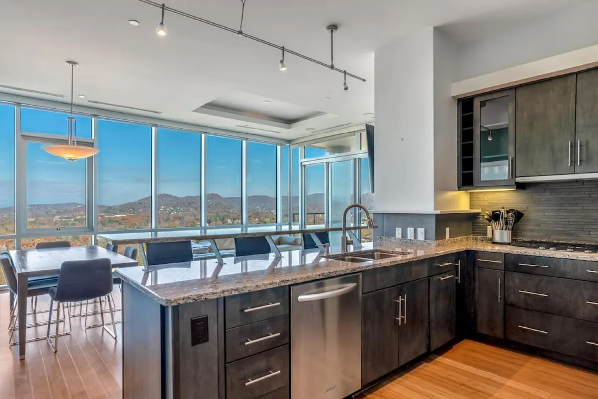 A stylish kitchen and dining area with a granite countertop, stainless steel appliances, large glass windows, and a dining area set against a scenic backdrop. Natural light fills the space, highlighting the modern design and ample room for meal preparation and casual gatherings.