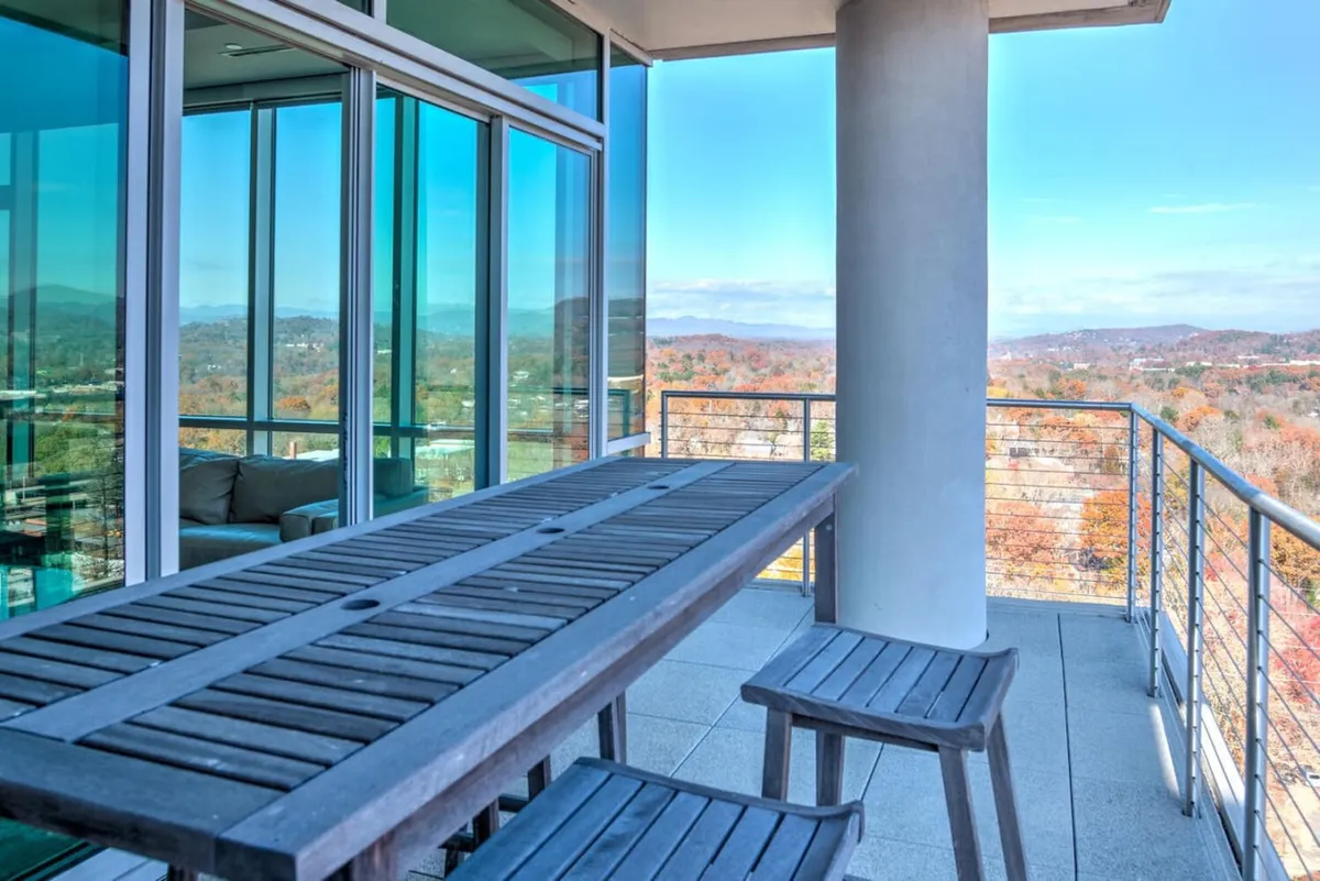 A tranquil exterior scene featuring a wooden table and stools on a terrace, framed by a sleek glass railing and a panoramic mountain view in the background. The mood is peaceful, highlighting opportunities for outdoor dining or simply soaking up the scenery.