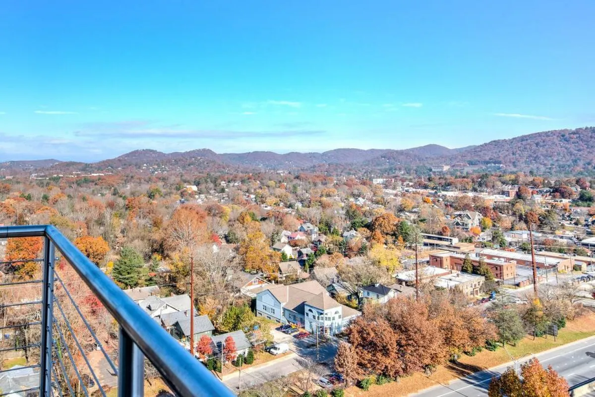 A sweeping view from an elevated vantage point, showcasing the urban landscape of Asheville nestled against the dramatic Blue Ridge Mountains. Vibrant autumn foliage adds warmth and color to the tranquil scene, while the soft evening light hints at a romantic sunset. The presence of a balcony suggests a private space to enjoy the panorama.