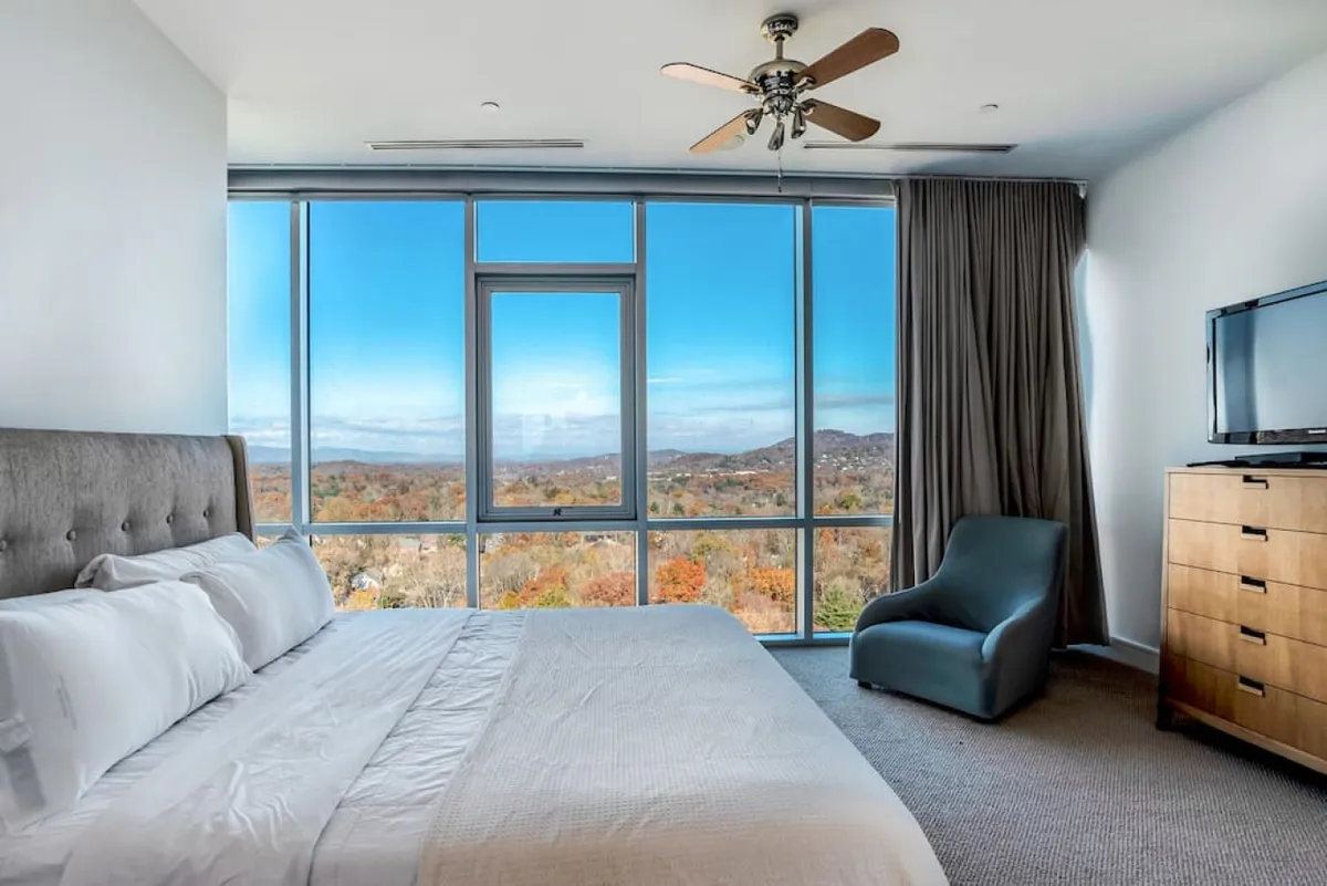 A comfortable bedroom with large windows overlooking scenic fall foliage, a bed adorned with plush pillows, a modern chair, and a wooden dresser. The ambiance is serene, with soft lighting encouraging relaxation and quiet moments.