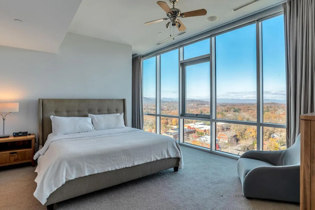 A serene bedroom featuring a plush bed positioned near expansive windows that frame breathtaking mountain views. The room is accented by a nightstand and a cozy armchair, ideal for relaxing or reading. Soft natural light and tranquil tones create a peaceful atmosphere.