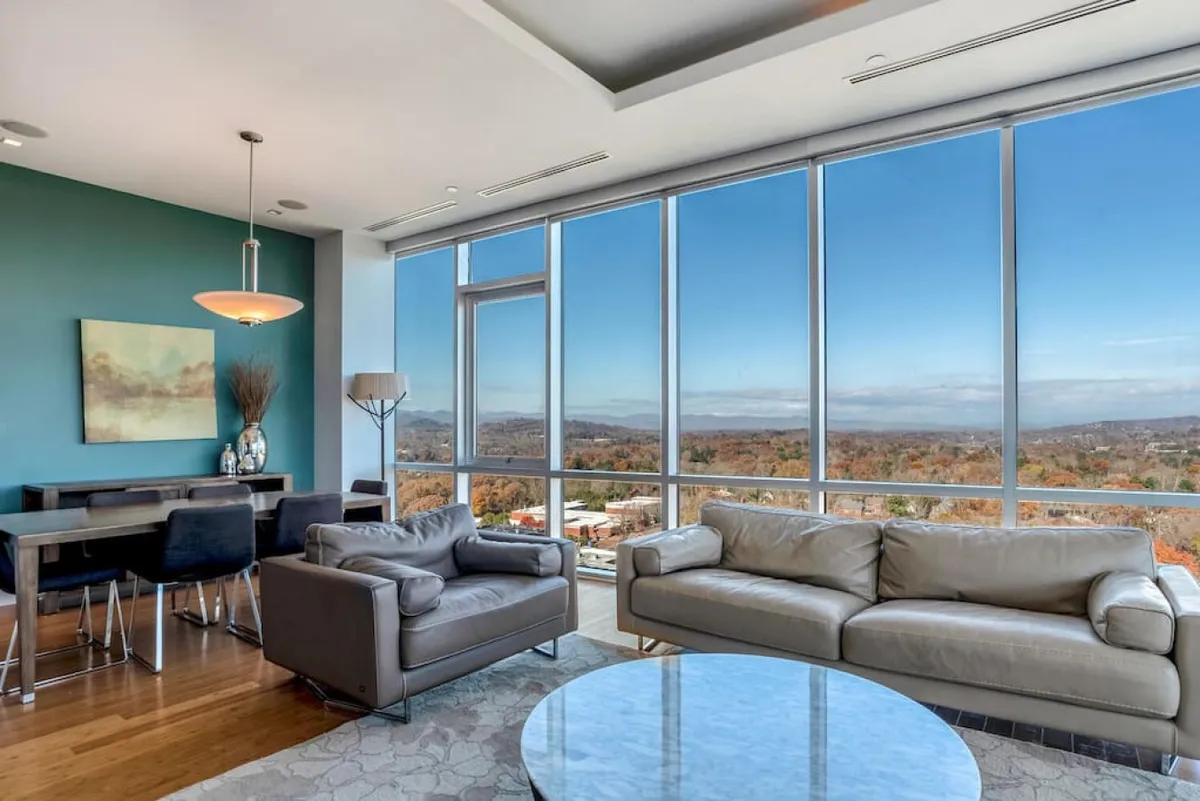 This image features a seamless living and dining area where oversized windows flood the space with natural light and reveal the stunning backdrop of mountain scenery. The stylish dining table is positioned for memorable meals with a view, complemented by comfortable seating and decorative modern artwork. The overall mood is tranquil and modern, perfect for intimate dinners or lively gatherings.