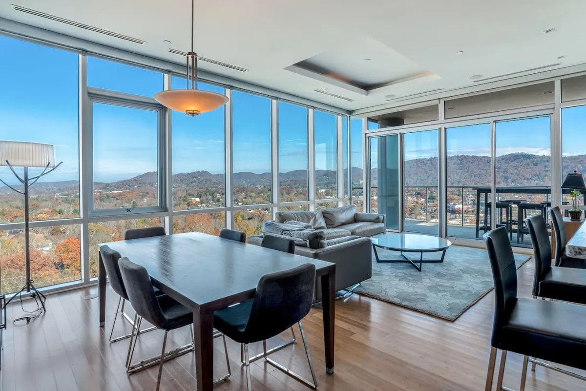 This image spotlights a contemporary dining area paired with a cozy living space, both oriented toward expansive windows framing the Blue Ridge Mountains. The scene is set for family gatherings or casual dining, with comfortable seating, elegant lighting fixtures, and modern decor completing the urban retreat feeling. Natural light pours in, and the setting encourages both celebration and relaxation.